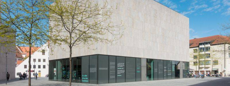 Besuch Jüdisches Museum München während der Klassenfahrt nach München. Auf dem Bild sehen wir den Beton-Glas-Kubus des Museums, davor zwie Bäume mit den ersten Frühlingsblättern.