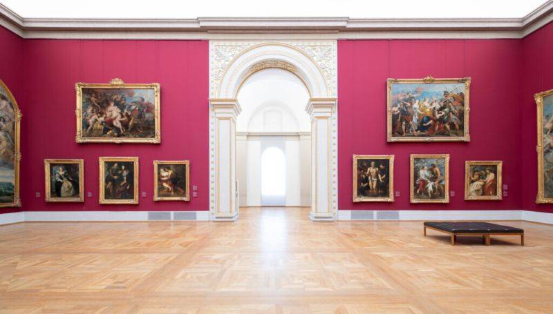 Während der Klassenfahrt nach München besucht die Schulklasse die Alte Pinakothek. Im Bild ein Ausstellungssaal. der Blick geht auf ein Durchgangsportal, rechts und links daneben an den roten Wänden hängen prachtvoll gerahmte Ölgemälde.